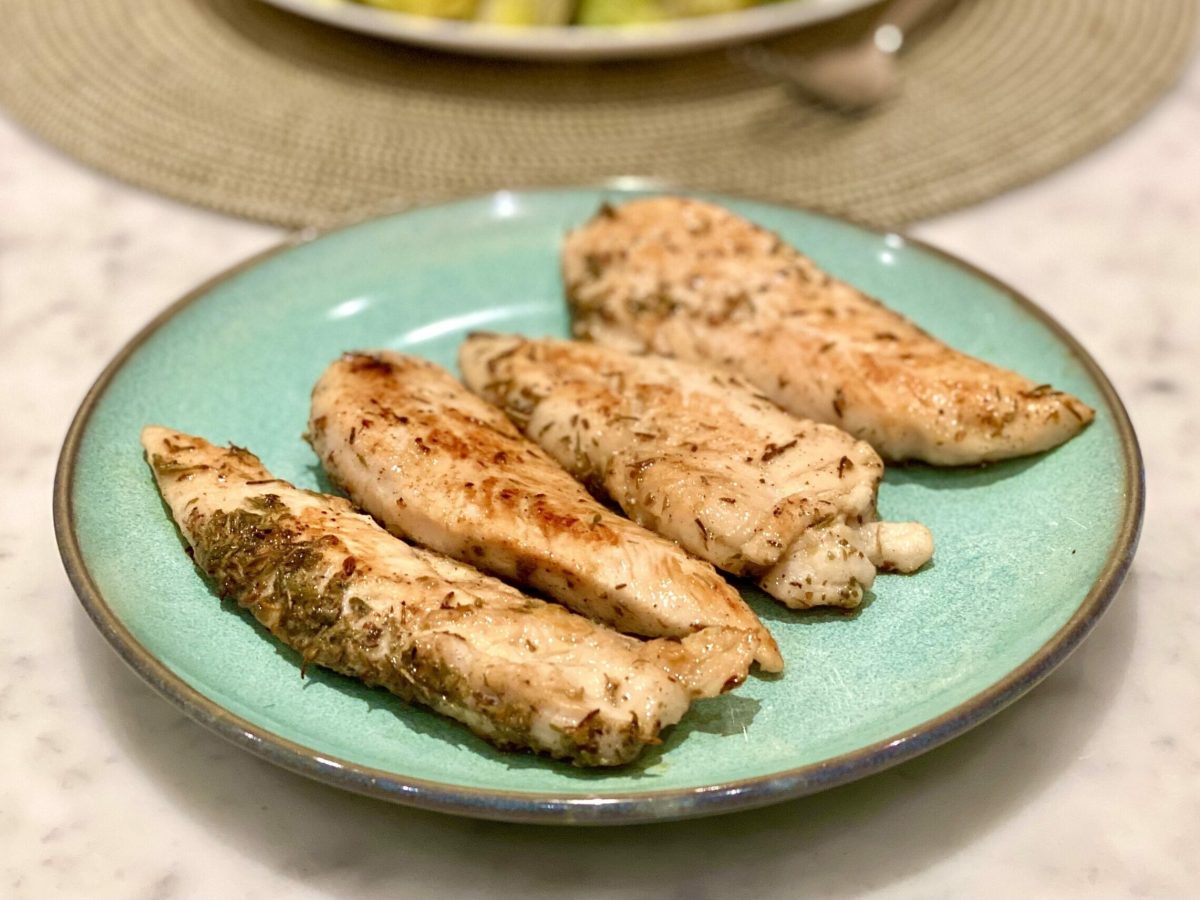 Four fillets of chicken, fried on a skillet with scattered herbs on top, served on a green plate. A recipe by Yalda Aloui.