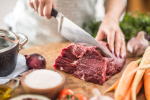 Meat on a wooden chopping board, next to carrots and salt. Hands are seen cutting the meat with a large knife.