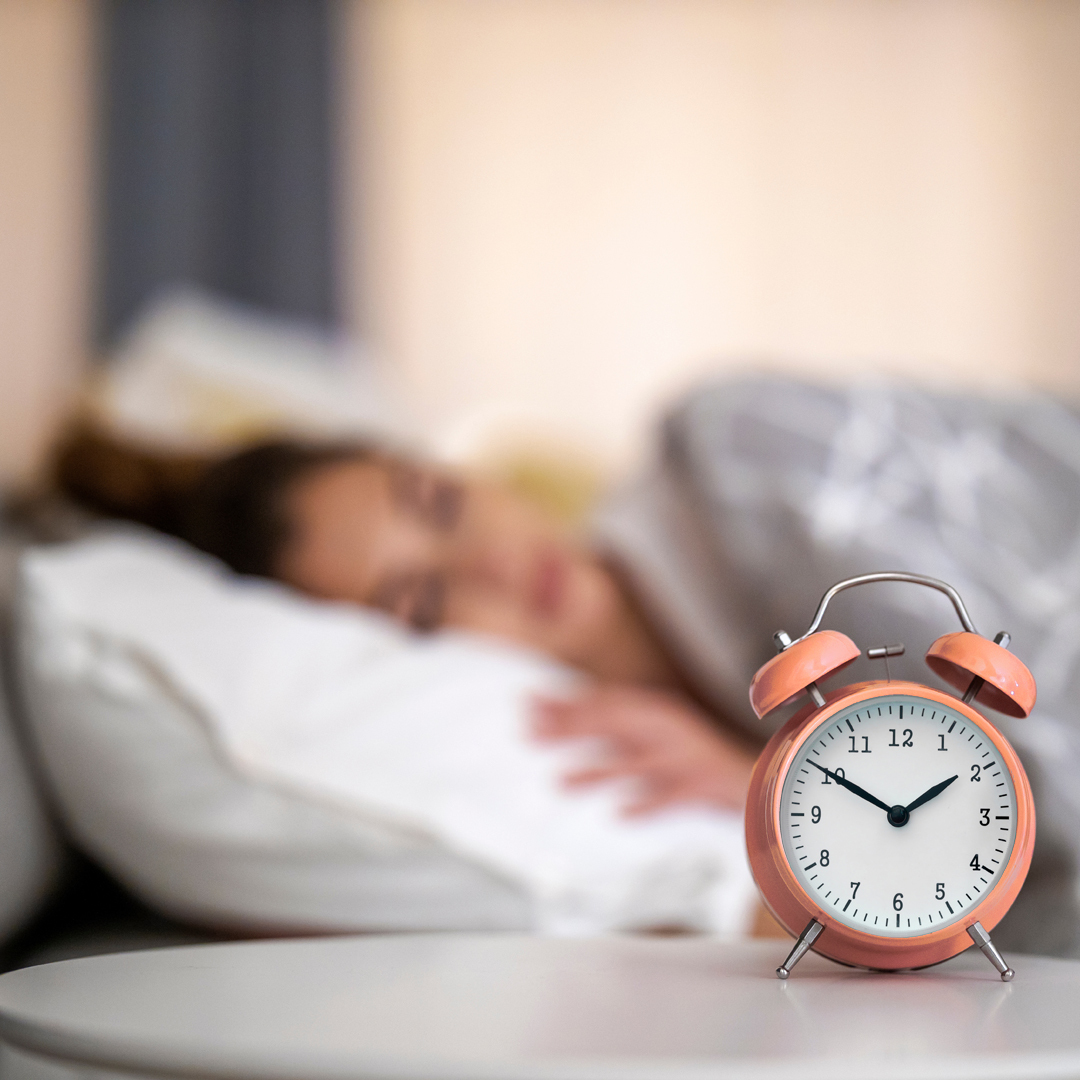 A salmon-coloured clock with at the forefront with a blurry picture of a female awake in bed.