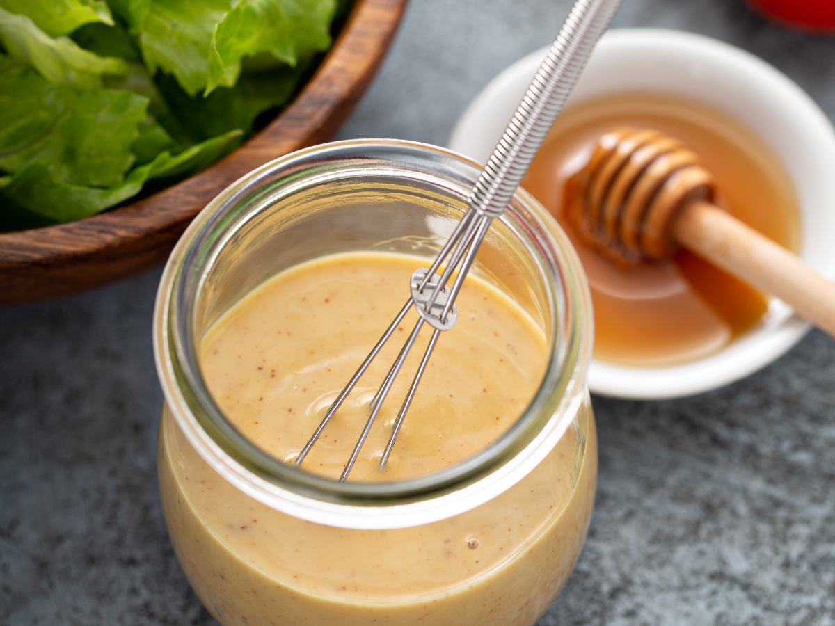 A glass jar of salad dressing with a bowl of honey at the side and a glimpse of lettuce in a bowl.
