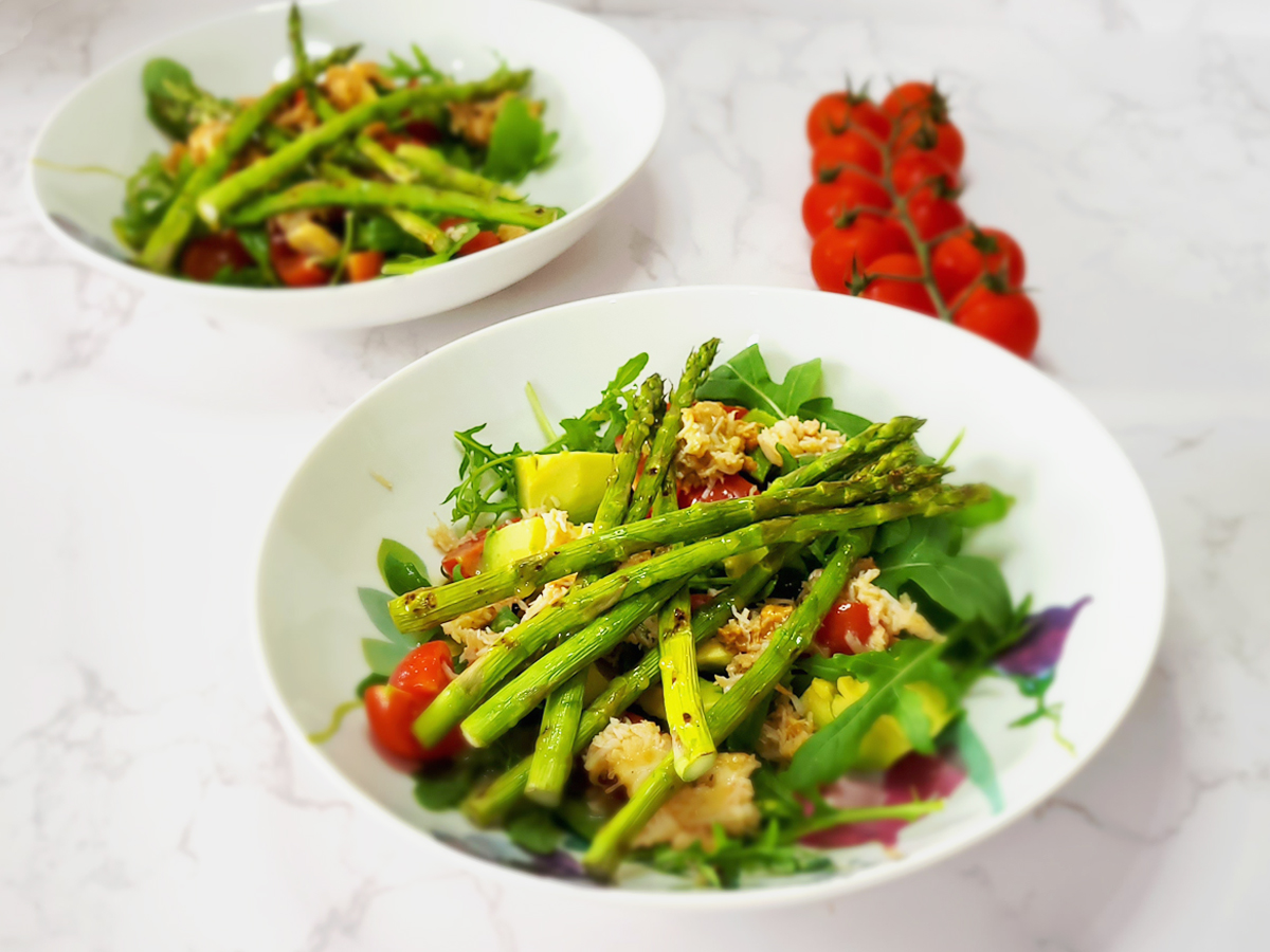 Two bowls with asparagus, crabmeat, arugula, tomatoes, and a dressing, with some cherry tomatoes by the side.