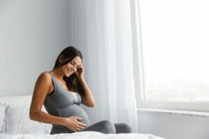 Female, pregnant, sat on her bed, smiling and holding her tummy.