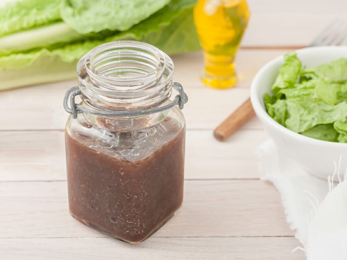 jar of balsamic dressing on a table surrounded by a bowl of salad and lettuce leaves