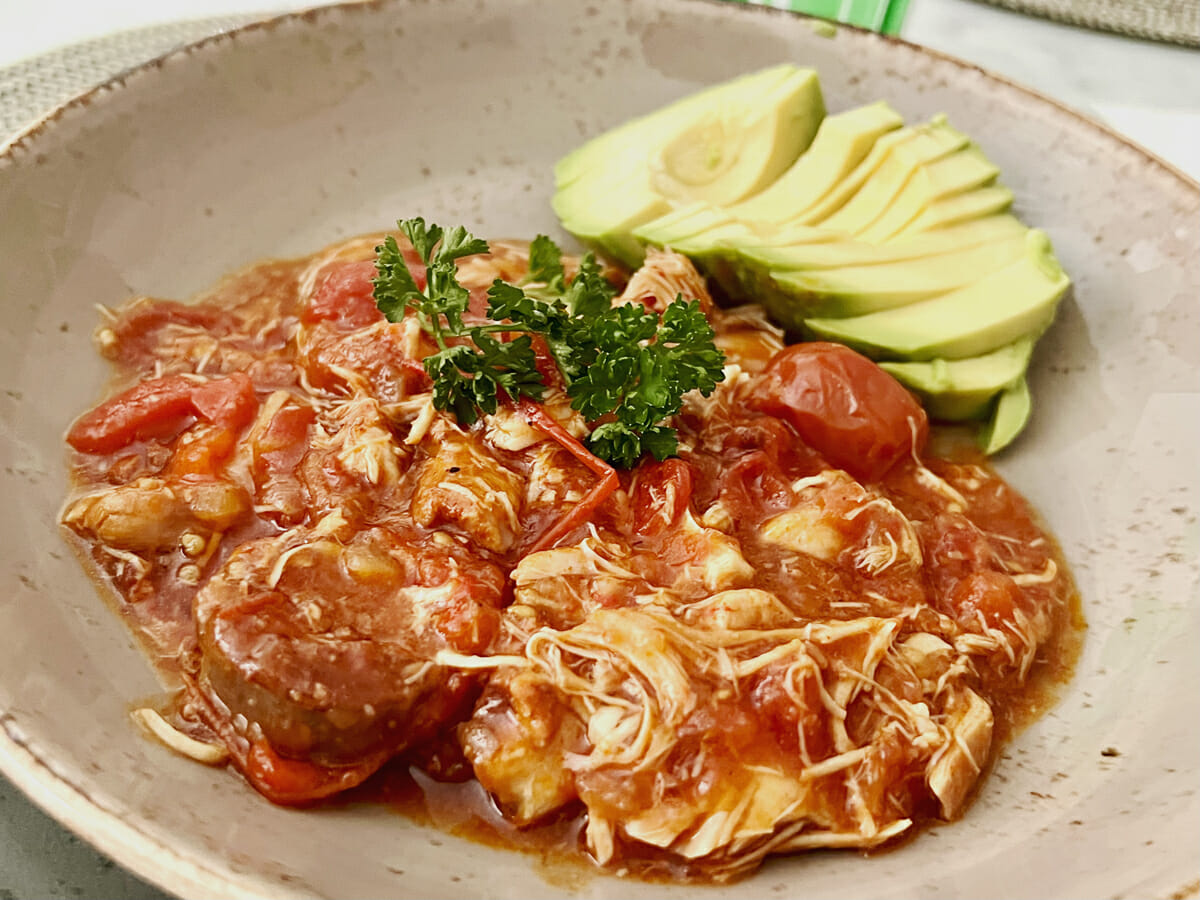 A bowl of chicken slow cooked in a red sauce served with sliced avocado on the slide and topped with a sprig of fresh parsley