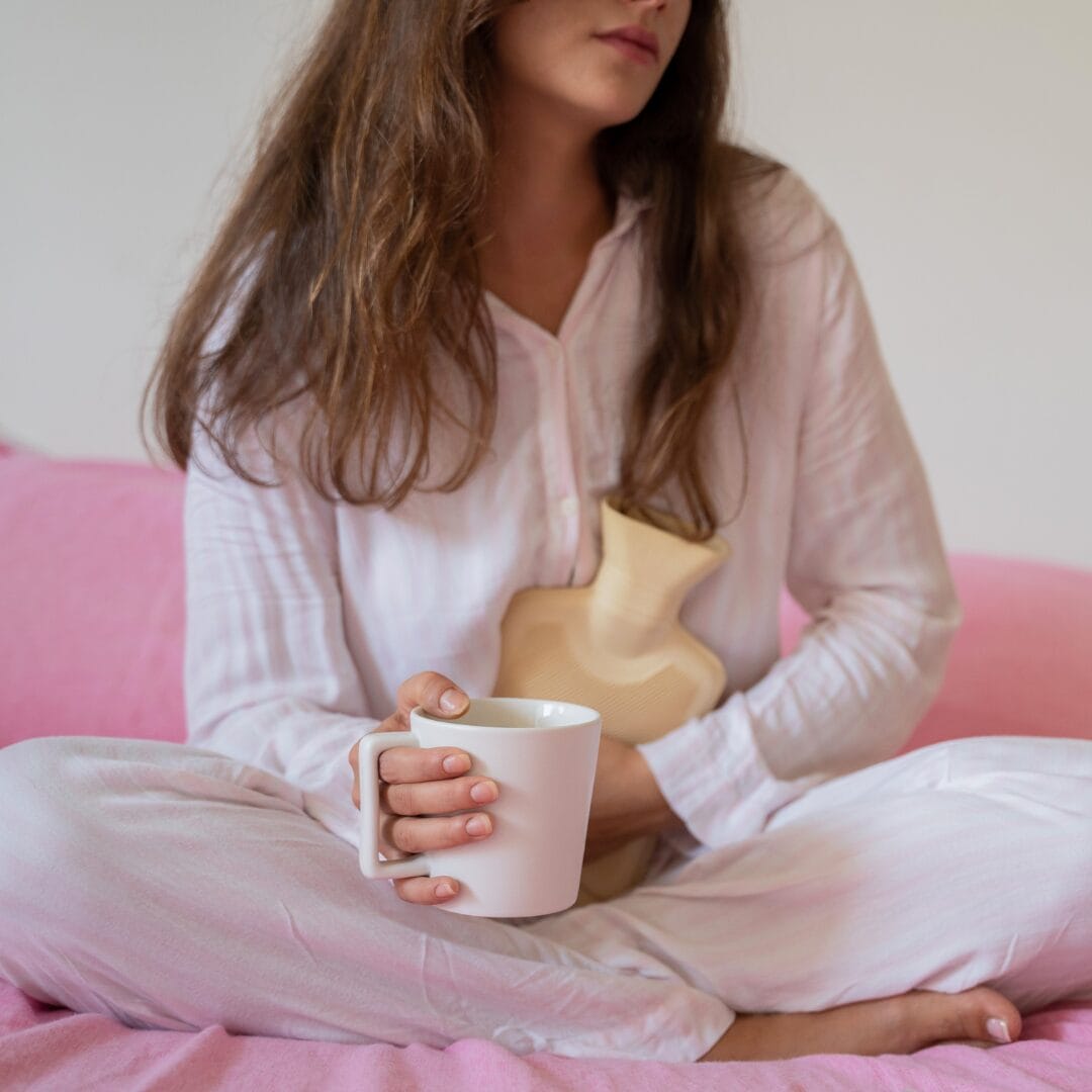 woman with pms symptoms clutching a hot water bottle and a hot drink