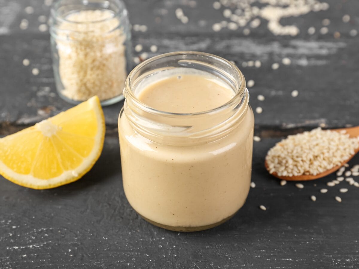 glass bottle with salad dressing made with lemon and tahini. Picture shows slice of lemon and tea spoon of scattered sesame seeds.